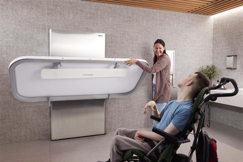 Person using a modern bathroom facility with a wheelchair and another person assisting.