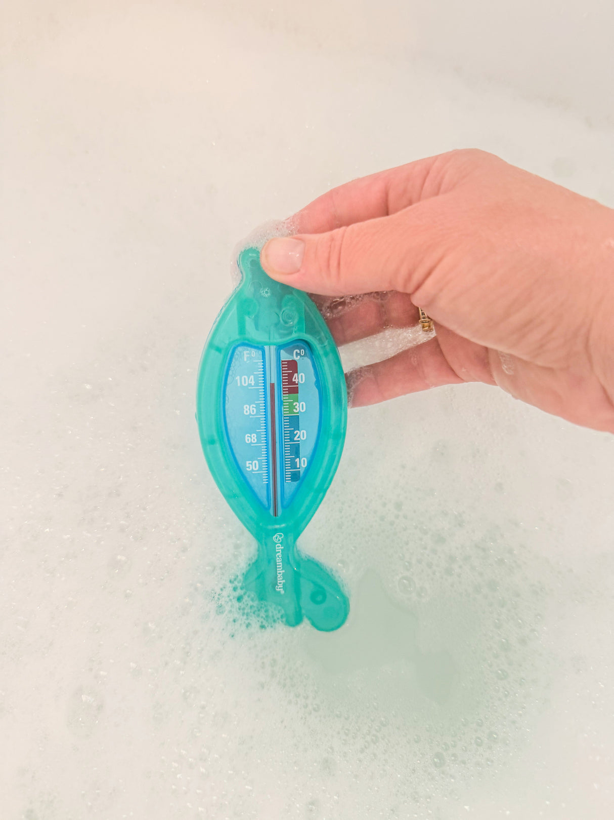 Hand holding a blue fish water temperature tester in a bathtub with bubbles.