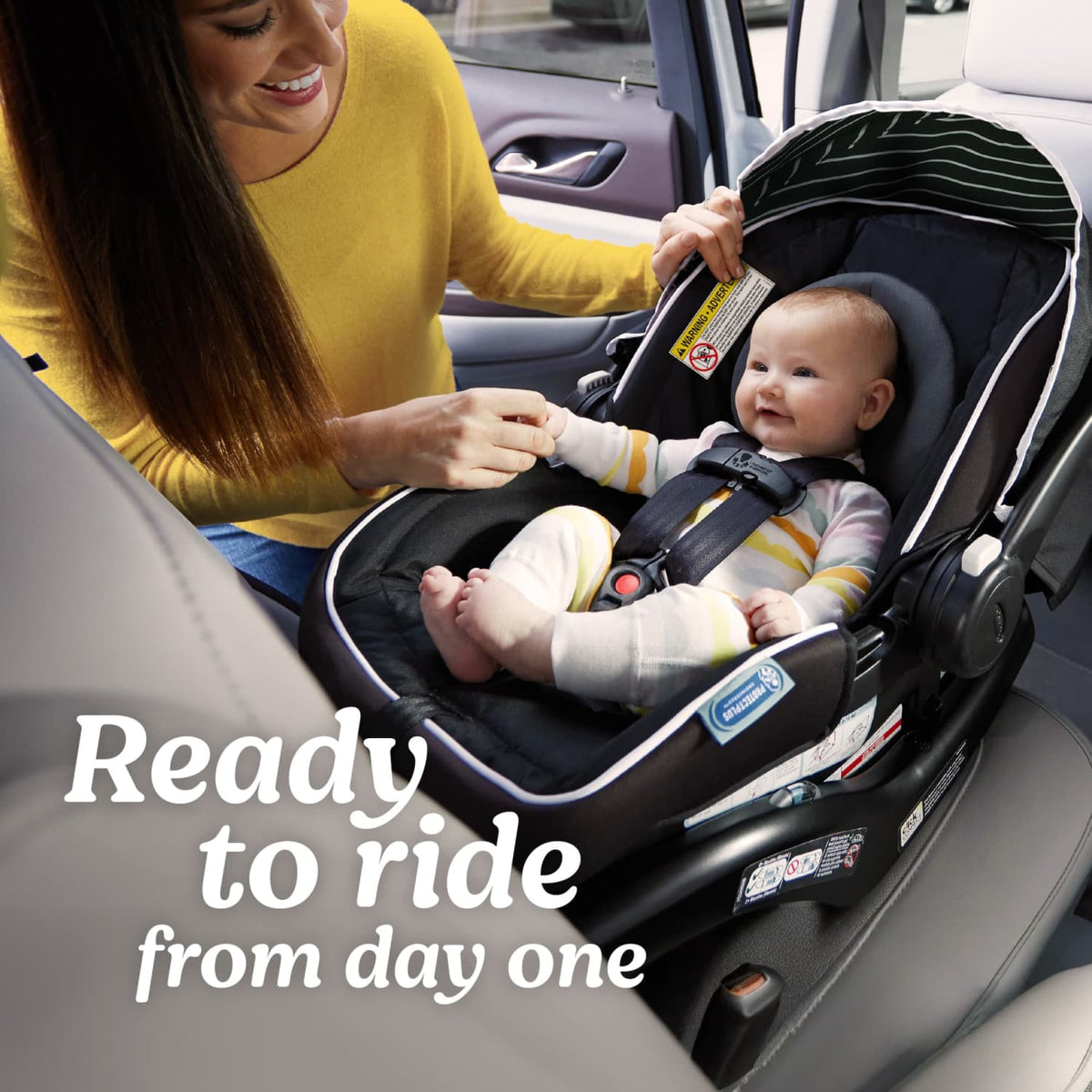 A image of a baby sitting in the Graco Snugride Lite LX Infant Car Seat installed in a car with a woman caretaker in a yellow shirt leaning over the seat. Graphic text on the image reads "Ready to ride from day one." 