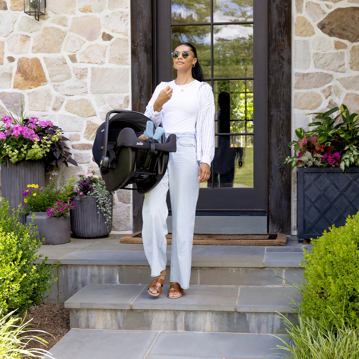 Woman in white outfit holding a baby car seat outside a stone building with plants around.